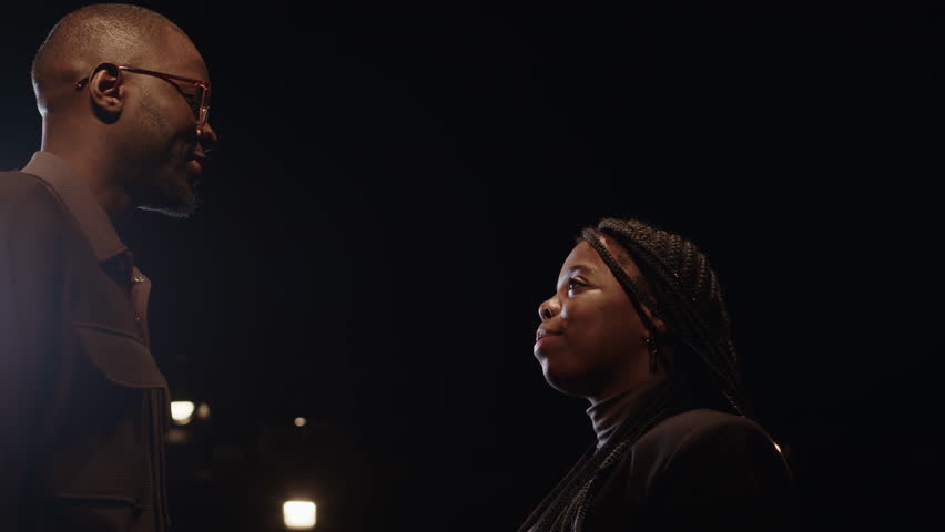 Low angle slowmo of young Black couple looking affectionately at each other while standing on dark stage under projector light during auditions for play at theatre Royalty-Free Stock Footage #3469022639