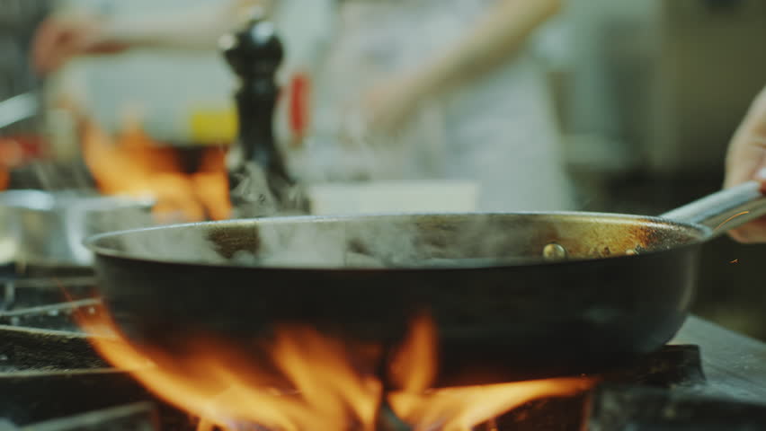 Close-up view of hands of master chef flipping food in frying pan as cooking on gas stove with burning fire flames in restaurant kitchen