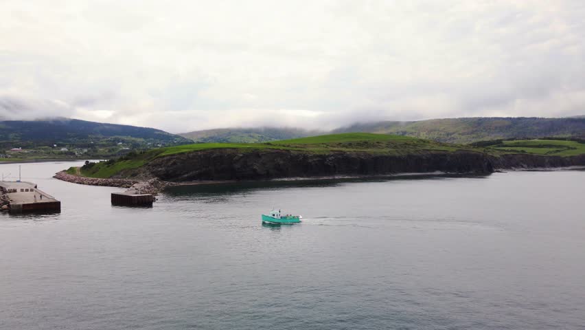 Drone shot of a small commercial lobster fishing boat coming back into the harbour after a long day of working revealing the beautiful landscape of the Cape Breton Coast in Nova Scotia, Canada.