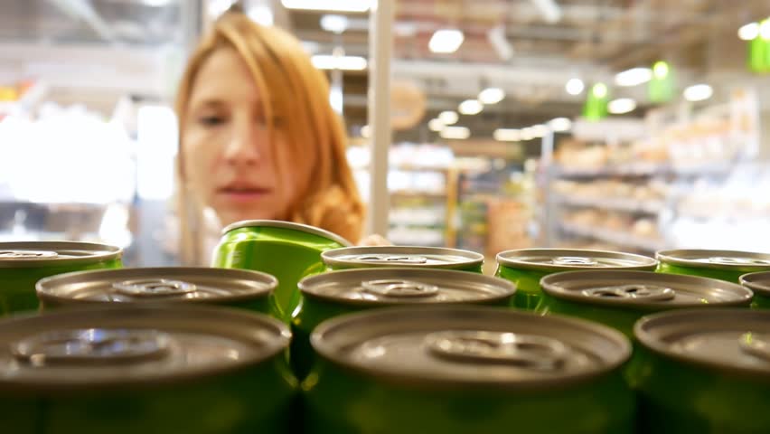 Close-up of many cans of soda in a supermarket fridge and cute girl opens the door and takes one
