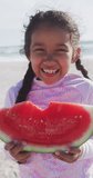 Vertical video of portrait of happy biracial girl eating watermelon on sunny beach. healthy, active time beach holiday.