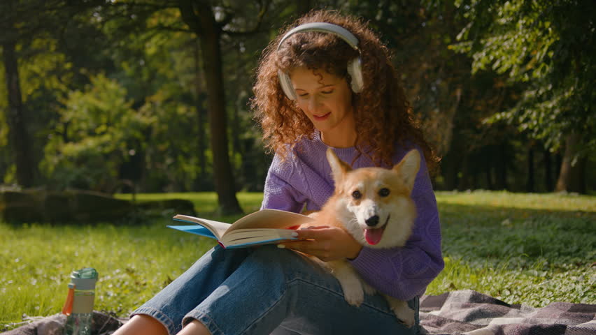 Caucasian woman young gen z student female domestic pet owner reading book on grass in city park outdoors smiling carefree girl in headphones rest on nature summer weekend with dog welsh corgi puppy Royalty-Free Stock Footage #3469173991