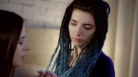 eccentric young woman with black and blue dreadlocks is sitting in a room and touching hand of her pretty girlfriend