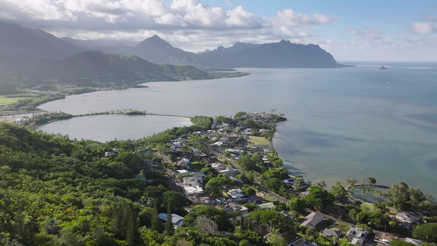Houses on green hill overlooking Kaneohe bay in blue ocean. Small countryside village with Kualoa ranch views. Sunny day Hawaii. Aerial Kahaluu residential community on windward coast of Oahu island