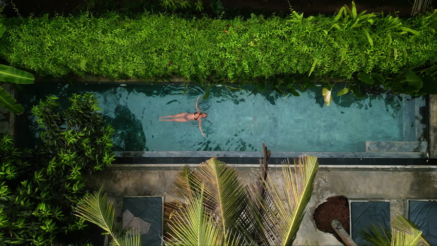 Overhead drone view of dark-skinned woman leisurely swimming in clear tropical pool surrounded by greenery. Peaceful vibe, perfect for wellness retreats, solo travel escapes, luxury resort promotions.