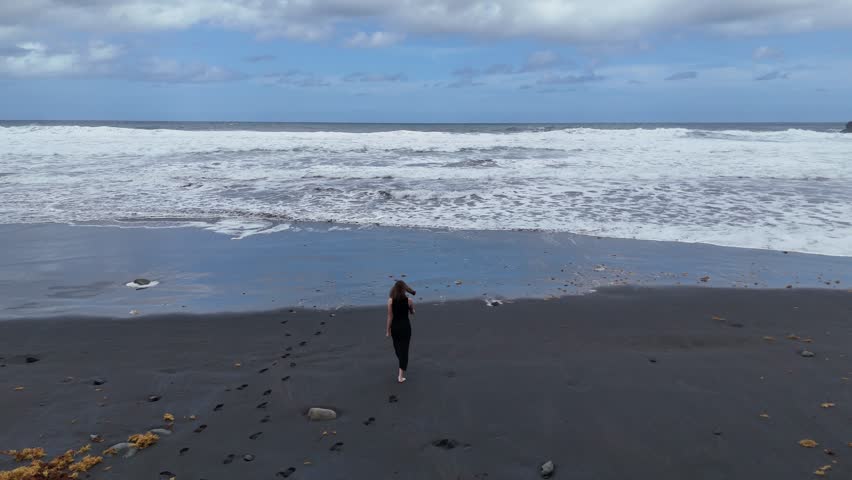 Aerial view of beautiful young woman walking on the sandy black beach in the black bikini near the sea with waves. Summer vacation on Canary island, Spain.