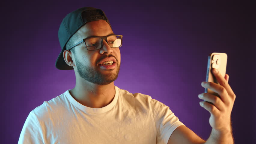A genial young man in a cap and glasses engages in a lively video call on his smartphone, gesturing and expressing himself enthusiastically against a violet backdrop.