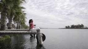 African woman sitting in a lake dock enjoying and connecting with nature. Serene footage scene of a female meditating front of a river, looking at the view and breathing. Spiritual retreat concept.