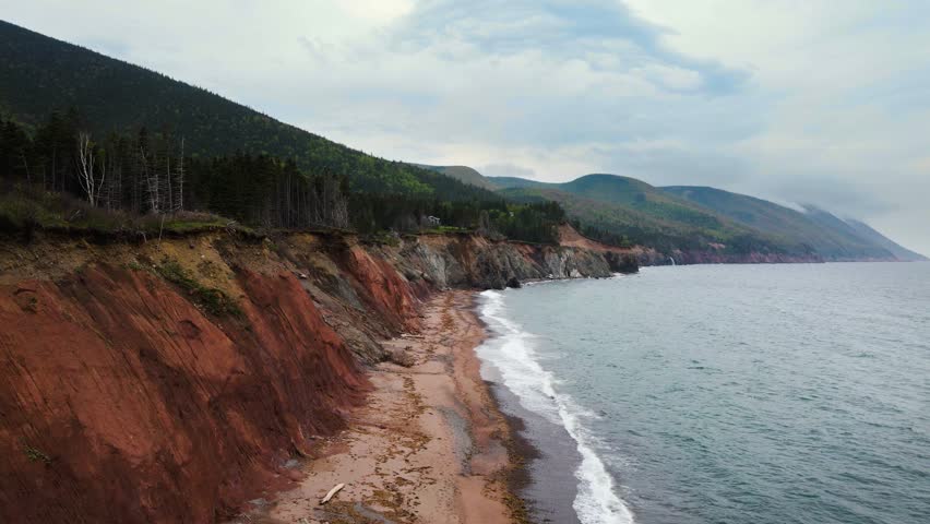 Drone shot revealing anisolated beach with a massive red cliff with mountains in the background and the Atlantic Ocean crashing onto the beach. Located on Cape Breton Island, Nova Scotia in Canada.