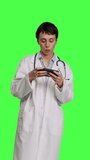 Front view General practitioner playing video games on mobile phone app, having fun with online gaming competition against greenscreen backdrop. Cheerful doctor relaxing with internet game. Camera B.