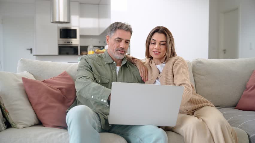 Happy middle aged older couple using laptop computer relaxing on couch at home. Smiling mature man and woman talking with device doing ecommerce shopping sitting on sofa in living room.