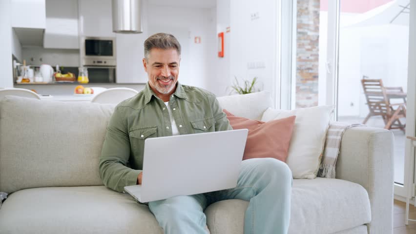 Happy middle aged man using laptop sitting on sofa at home. Mature older user looking at computer browsing internet, doing ecommerce shopping on website relaxing on couch in modern living room.