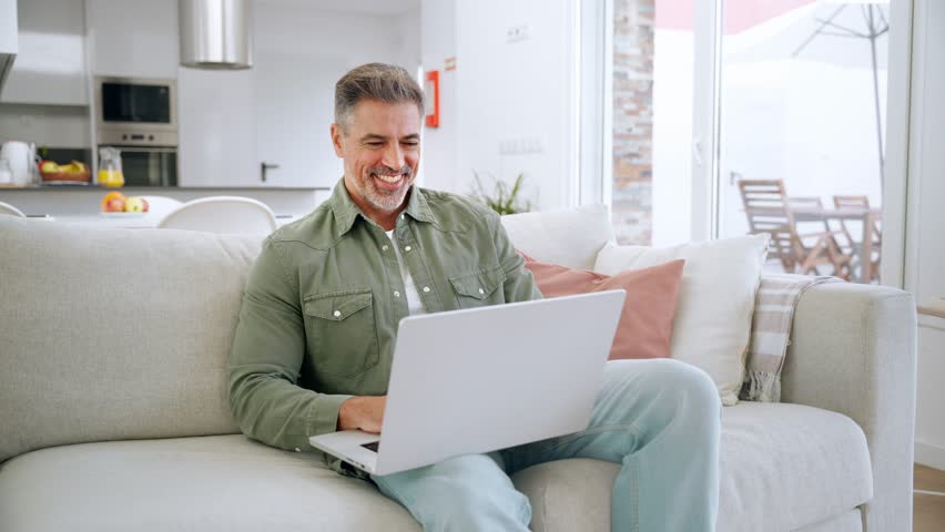 Happy middle aged man using laptop sitting on sofa at home. Mature older user looking at computer browsing internet, doing ecommerce shopping on website relaxing on couch in modern living room. - Powered by Shutterstock - Get 15% off with code: PIKWIZARD15