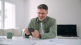 Smiling senior mature middle aged man wearing eyeglasses holding cell phone using smartphone sitting at home table, scrolling social media, checking financial apps, buying online, texting messages. - Powered by Shutterstock - Get 15% off with code: PIKWIZARD15
