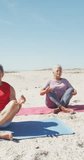 Vertical video of happy senior biracial couple meditating on sunny beach. healthy, active retirement beach holiday.