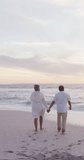 Vertical video of happy senior biracial couple walking on sunny beach. healthy, active retirement beach holiday.