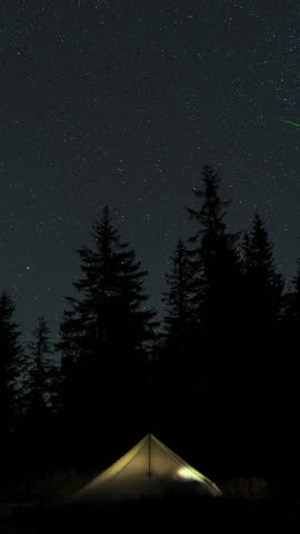 Time lapse of moving stars in the night sky above a glowing tent in the forest. Epic video, vertical footage