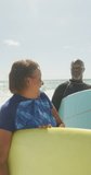 Vertical video of happy senior african american couple with surfboards on sunny beach. spending free time at beach holiday.