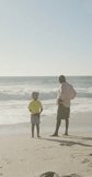 Vertical video of happy senior african american grandfather with grandson on sunny beach. spending free time at beach holiday.
