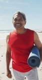 Vertical video of portrait of happy biracial man with yoga mat on sunny beach. healthy, active retirement beach holiday.
