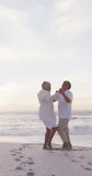 Vertical video of happy senior biracial couple dancing on sunny beach. healthy, active retirement beach holiday.