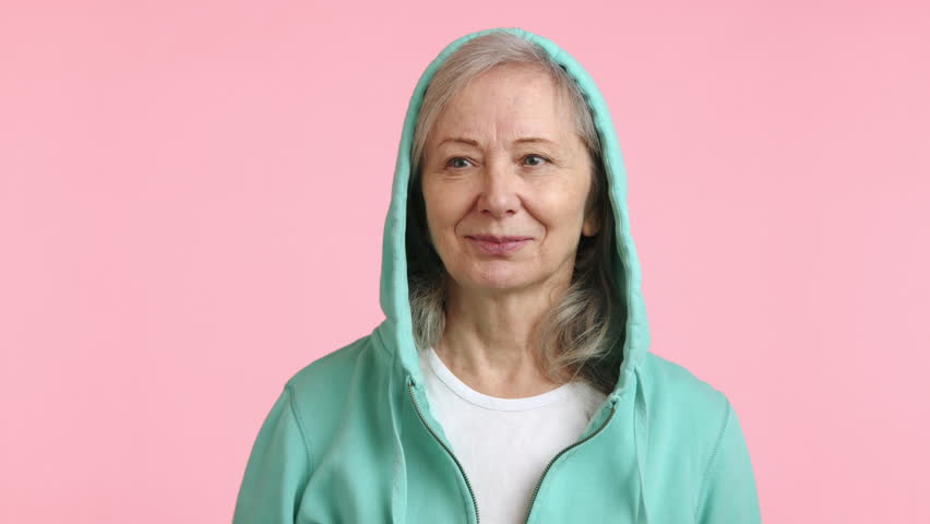 A senior woman with a searching gaze peers into the distance, hand to forehead, looking for someone or something that's not immediately visible, set against a soft pink background. Camera 8K RAW.  Royalty-Free Stock Footage #3469675019