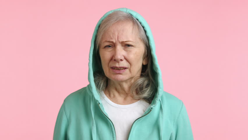 A distressed senior woman is covering her ears with her hands, showing a troubled reaction to a very loud and discomforting noise against a pale pink background. Camera 8K RAW.  Royalty-Free Stock Footage #3469676155