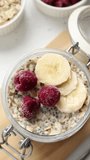 Hand sprinkling chia seeds on top of overnight oats (cold oatmeal) in a jar with bananas and raspberries. Top table view. Close-up. Preparation of healthy vegan plant-based meal for breakfast.