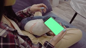 young woman using smartphone with green screen mockup in home living room to watch videos while lying on sofa, High quality 4K footage