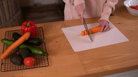 Close-up video of a young woman preparing food in a rustic kitchen, she is carefully chopping a carrot using a sharp knife on a chopping board placed over a hard wood kitchen top.

