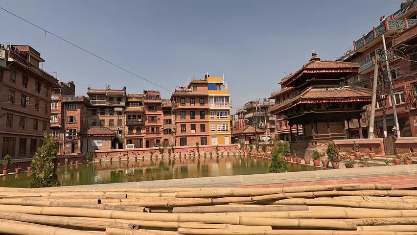 Bamboo logs stored at the vibrant green Bholachhe Pond in the core of Bhaktapur, Nepal. Plant pots and brick buildings surrounding the lake.