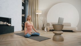 Relaxed blond hair woman sitting cross legged on yoga mat in modern living room, female fitness coach doing breathing exercise, yoga online, laptop video call, remote training at home, dolly wide shot
