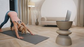 Sporty woman does stretching yoga exercises on mat in modern living room, female fitness coach practicing warrior position asana, online laptop video call, remote training at home, dolly wide shot