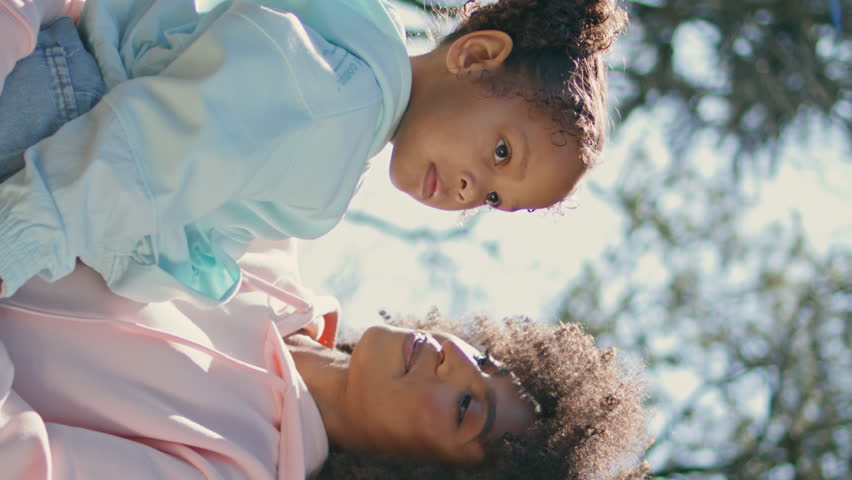 African american mom holding daughter standing on park alley close up. Cheerful smiling woman talking with adorable curly child at weekend walk vertically shot. Little cutie enjoying sunny holiday.