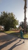 An enchanting young blonde woman in green outfit runs through a city palm park on an early summer morning. The camera moves towards her, capturing the scene in slow motion. vertical video