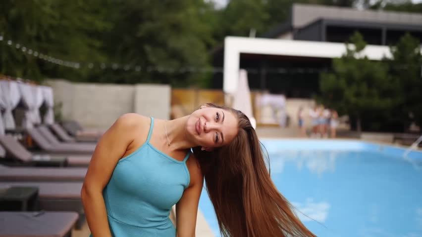 Pretty woman poses near swimming pool and sun loungers in blue dress, shows long hair and charming smile. Female model at summer pool party dancing and sending air kiss in club on holidays. Royalty-Free Stock Footage #3469902377