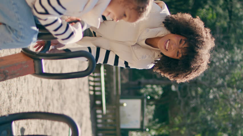 Cute child spinning at carousel in playground with cheerful mother. Happy african american girl sitting on merry-go-round having fun vertically view. Smiling woman helping daughter. Active childhood.