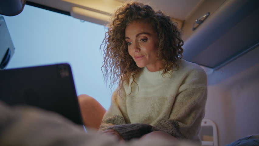 Woman traveller reading email on laptop sitting evening motorhome close up. Smiling curly lady looking at computer screen on trailer bed. Middle-aged businesswoman working online at camper weekend.