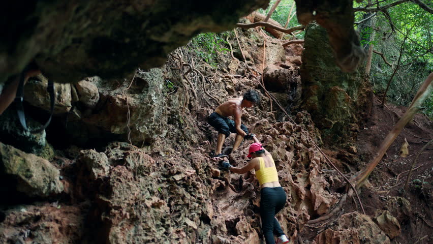 Asian man and woman climber helping each other during climbing on rocky mountain at tropical beach island. People enjoy outdoor active lifestyle extreme sports climbing on summer holiday vacation.