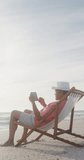 Vertical video of biracial man alone on sunbed on sunny beach. healthy, active retirement beach holiday.