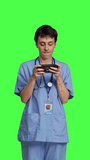 Front view Medical assistant playing mobile video games on smartphone app, taking a break from healthcare work. Nurse enjoys gaming tournament on phone, standing against greenscreen backdrop. Camera B