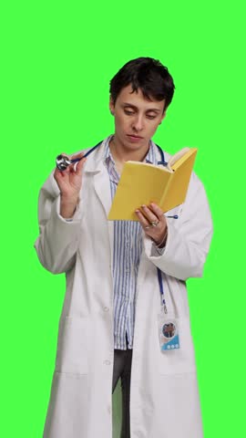 Front view Medic specialist reading a healthcare literature book to develop her medical expertise in the industry, stands against greenscreen backdrop. Clever doctor in white coat enjoys lecture Royalty-Free Stock Footage #3469975235