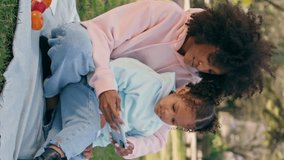Mother daughter looking at smartphone sitting blanket outdoors. Happy african american woman showing cartoon to little girl at family picnic vertical video. Smiling mom entertaining child with phone