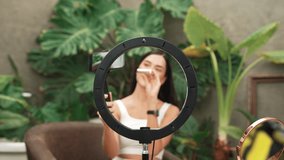 Video zoom in through center of circular ring light revealing woman use selfie stick for beauty and cosmetic tutorial video content. Beauty blogger amidst serene ambiance of natural garden. Blithe