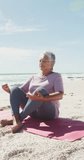 Vertical video of happy senior biracial woman meditating on sunny beach. healthy, active retirement beach holiday.