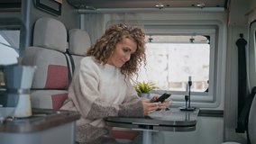 Happy freelancer reading sms smartphone sitting table trailer close up. Smiling curly woman looking cellphone screen relaxing in campervan. Carefree lady watching video content at mobile phone in van.