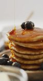 Vertical video of maple syrup pouring on stack of american style pancakes topped with blueberries. home cooking sweet treat, food, flavour and nutrition.