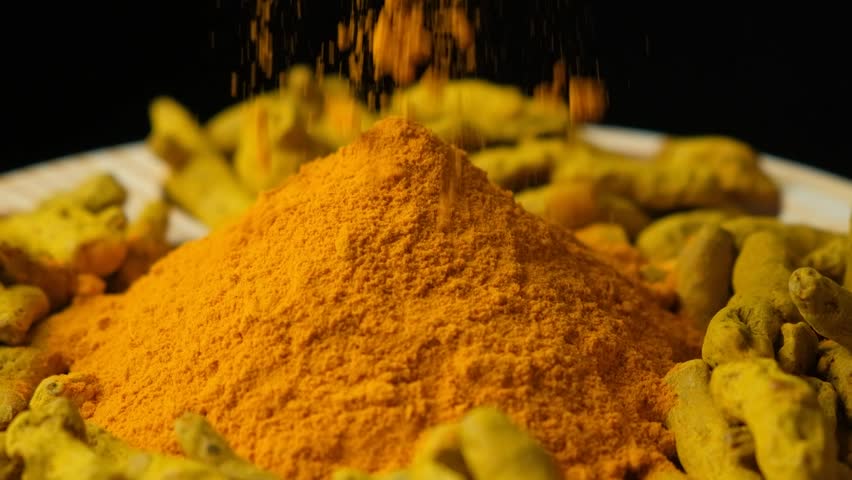 Turmeric powder sprinkled on wooden dish with powder and dry turmeric root, slow motion.