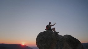 Happy backpacker shooting video on mobile phone sitting on the top of mountain in Yosemite national park, tiny orange sun hiding behind distant mountain. High quality 4k footage