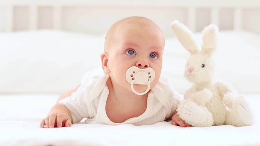 a baby sucks a pacifier on a bed in a white bodysuit in a bright bedroom, a cute baby with blue eyes lies on his stomach with a pacifier in his mouth and a plush bunny Royalty-Free Stock Footage #3470244095